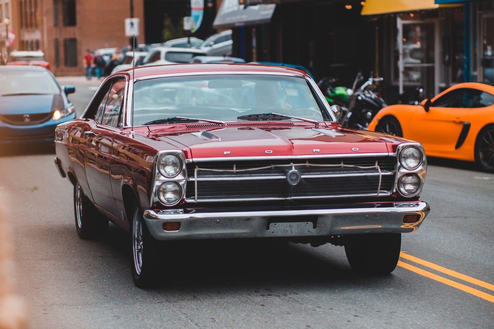 A red 1966 Ford Fairlane photographed on a town street, with buildings and parked cars behind it