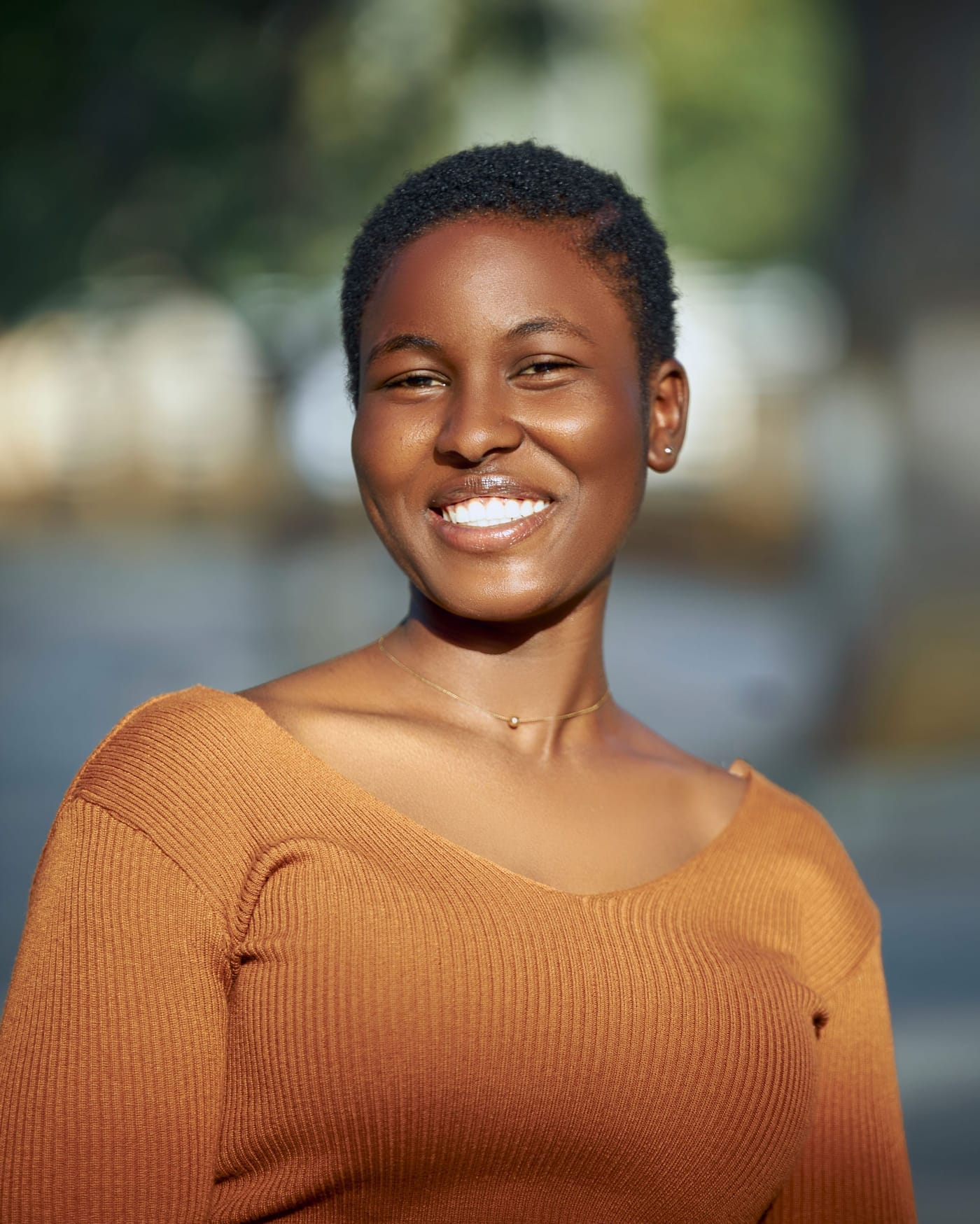A smiling woman in a rust-orange knit top, photographed outdoors against blurred greenery